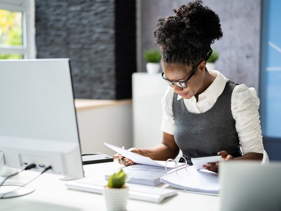 Eine Frau sitzt am Schreitische uns arbeitet mit einem Aktenorder